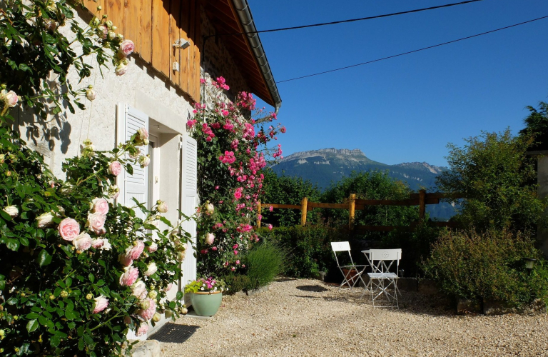 La faade d'une maison abrite baigne dans un soleil radieux. La porte d'entre, cerne de rosiers, ouvre sur une alle o se trouvent une table et deux chaises positionnes prs d'arbustes, orientes vers une montagne et son ciel bleu.