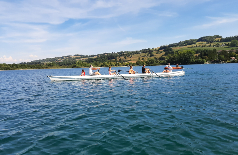 Sur les eaux calmes du lac, un groupe de personnes rame  bord d'un long aviron. En dernire position, une enfant admire le paysage.