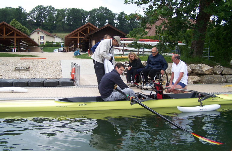 Depuis un quai d'embarquement, une personne en fauteuil roulant, encadre par des moniteurs et ses accompagnateurs, s'apprte  embarquer  bord d'un aviron adapt pour l'accueillir en pratique double.
